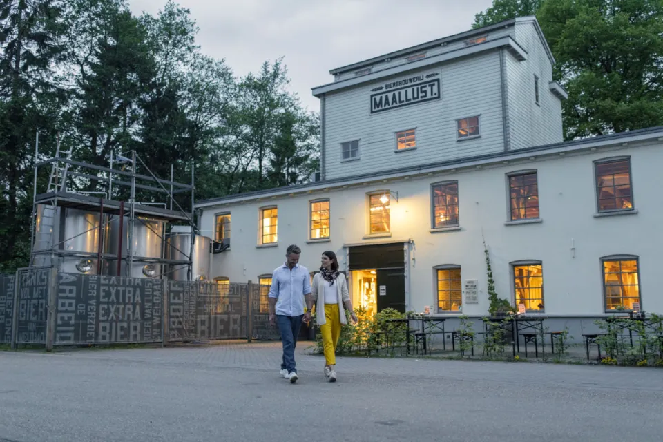 Verlicht wit gebouw van brouwerij Maallust in Veenhuizen met bezoekers op het plein