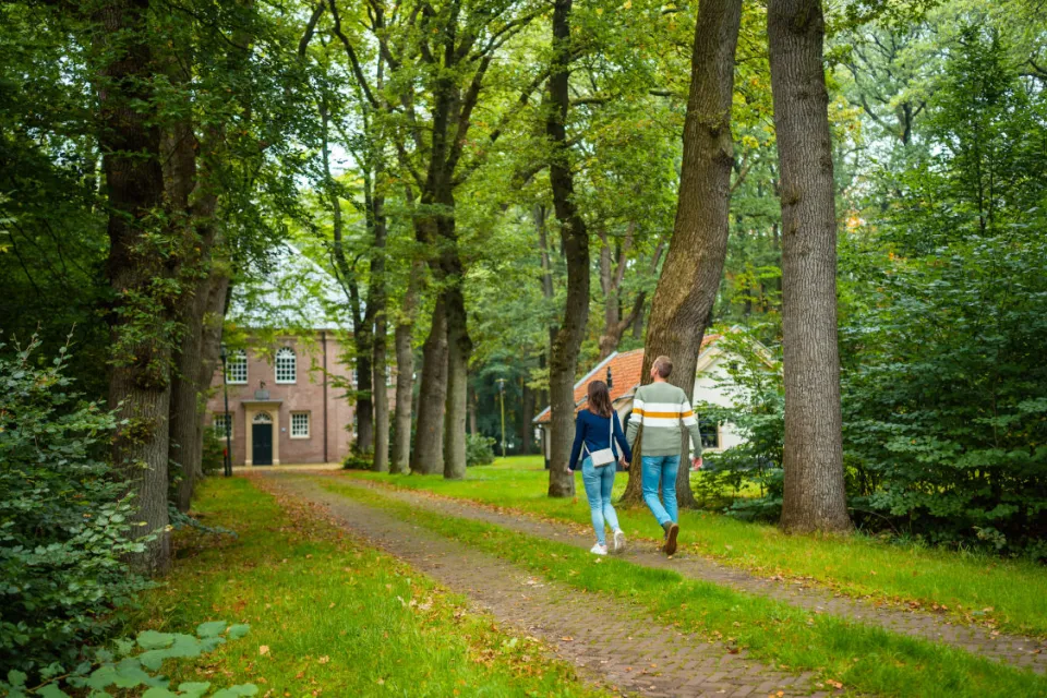Twee wandelaars lopen over een bospad langs een historisch gebouw in de Kolonien van Weldadigheid in Drenthe