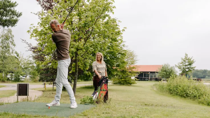 Twee gasten spelen golf op de baan van golfpark Hildenberg met het clubgebouw op de achtergrond